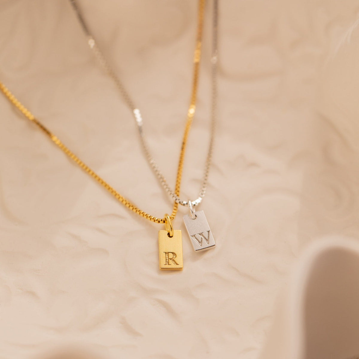 Two Mini Initial Tag Necklaces—a gold one with an "R" and a silver one with a "W"—rest on a textured surface. These personalized necklaces make a thoughtful Christmas gift and are perfect for adding custom style.