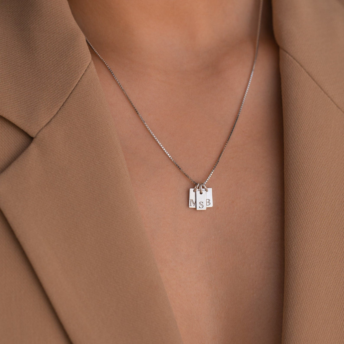 A close-up of the Mini Initial Tag Necklace with N, S, and B letter pendants, paired with a tan blazer—an elegant personalized accessory and an ideal Christmas gift.
