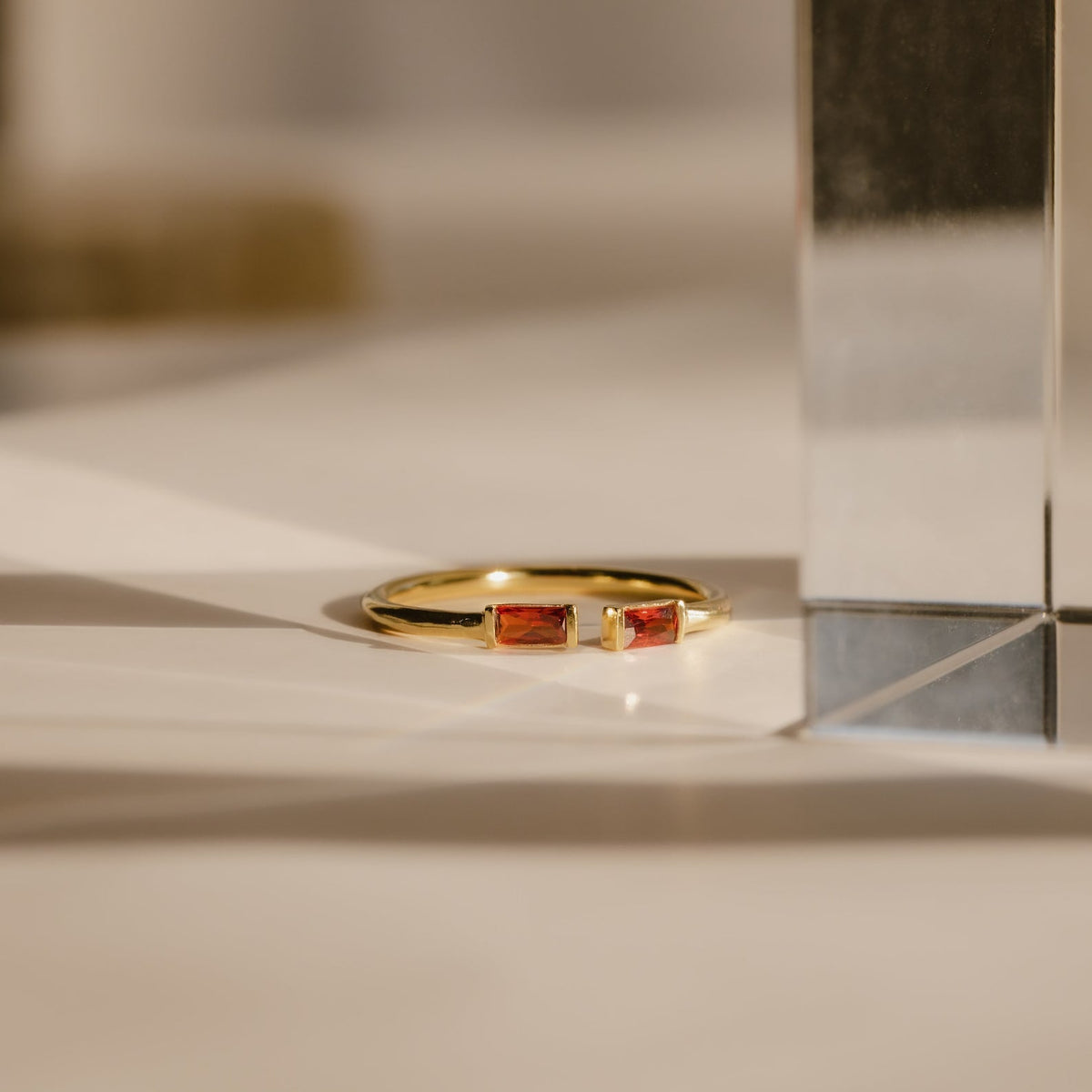 The Duo Baguette Birthstone Ring, featuring two rectangular red stones, rests on a white surface in gentle sunlight—a beautiful choice for a personalized gift.