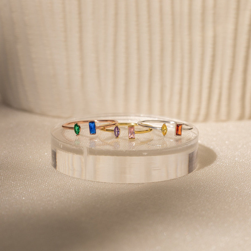 Five handcrafted rings with vibrant rectangular gemstones are displayed on a clear stand in soft light, highlighting the Duo Marquise Baguette Ring for a personalized gemstone jewelry touch.