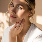 A woman with blonde hair in a white top gently touches her face, softly lit and wearing gold jewelry, including the Duo Marquise Baguette Ring.