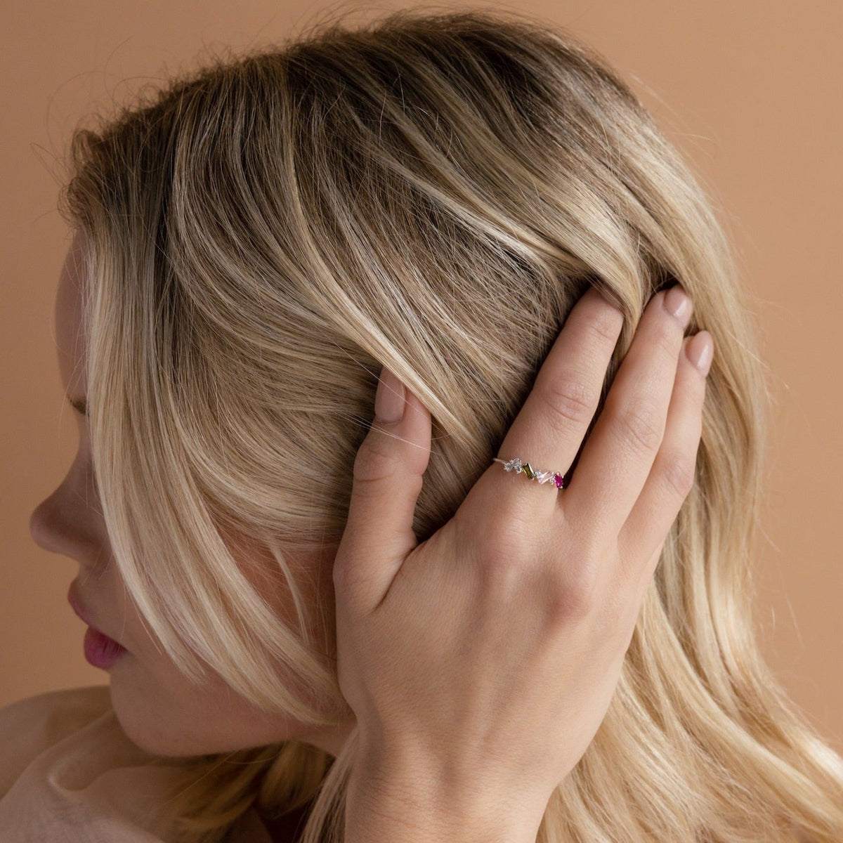 A woman with blonde hair touches her head, showcasing the Cluster Baguette Birthstone Ring in Rose Gold adorned with vibrant gemstones on her finger.