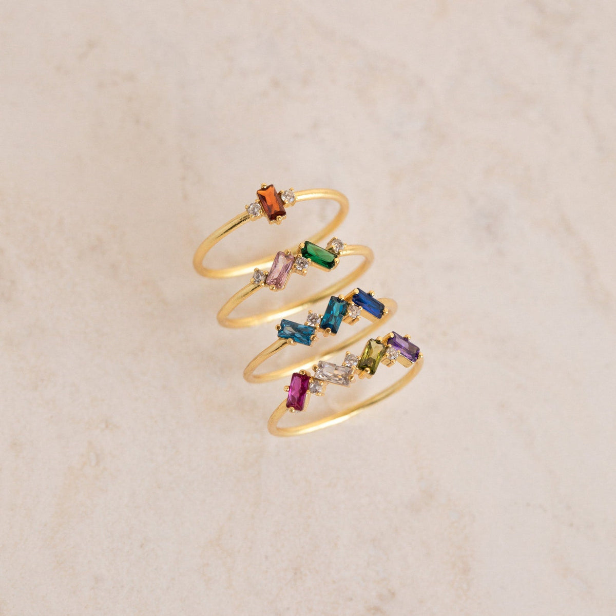 Four Cluster Baguette Birthstone Rings, each featuring colorful rectangular gemstones, are arranged in a spiral on a light marble surface—perfect as personalized statement or unique Mother's rings.