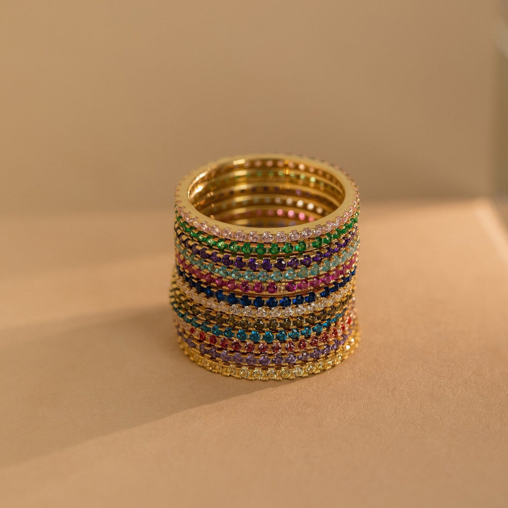A stack of Eternity Birthstone Rings is set with colorful gemstones and arranged on a beige surface under warm lighting—an elegant, customizable ring and the perfect Christmas gift.