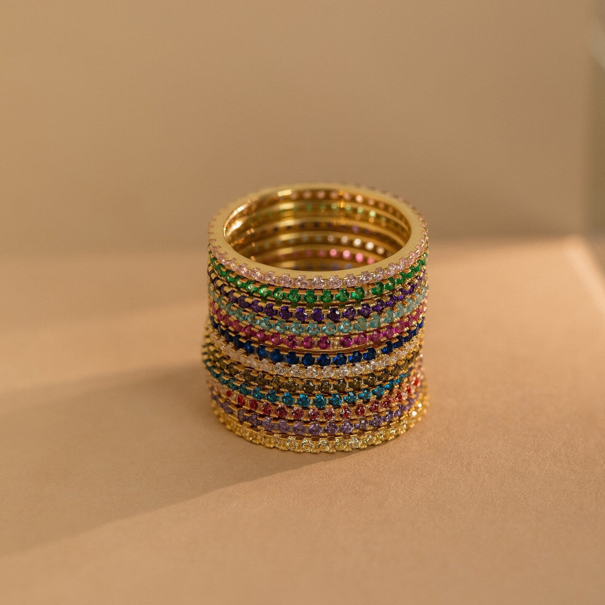 A stack of Eternity Birthstone Rings is set with colorful gemstones and arranged on a beige surface under warm lighting—an elegant, customizable ring and the perfect Christmas gift.