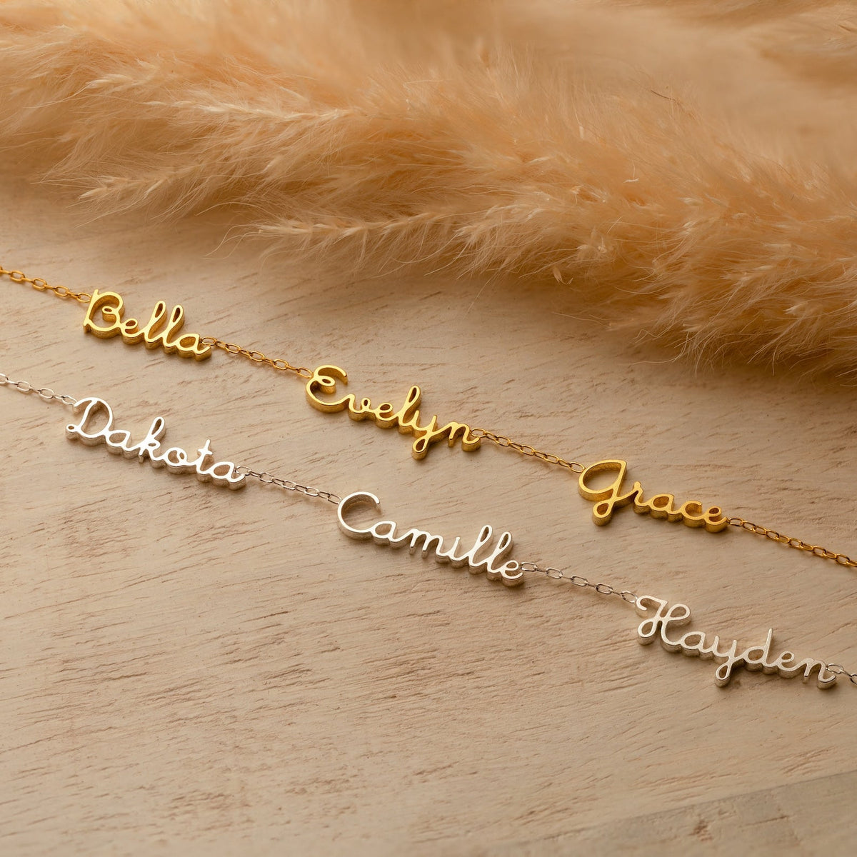 Three gold and three silver Dainty Tiny Multiple Name Necklaces with personalized nameplates are displayed on a light wooden surface beside brown pampas grass.
