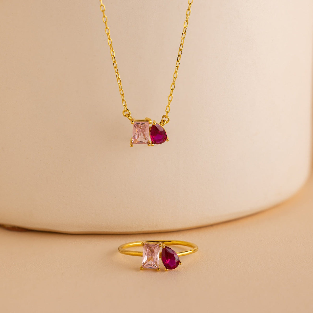 A gold necklace and the Toi et Moi Birthstone Ring in Rose Gold, featuring pink square and red teardrop gems, elegantly displayed on beige—a customizable ring that makes a thoughtful Christmas gift.