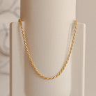 Gold rope chain necklace hanging in a graceful arc on a minimal cream ceramic vase.