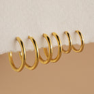 Six gold Mini Bold Hoops from our collection hang on the edge of a white textured surface, showcasing their varying sizes and timeless style.