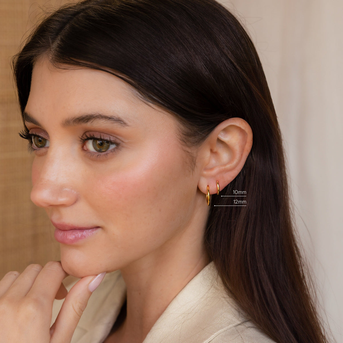 A woman with long brown hair wears two small gold hoop earrings from our Mini Bold Hoops Set, labeled 10mm and 12mm, on her left ear.