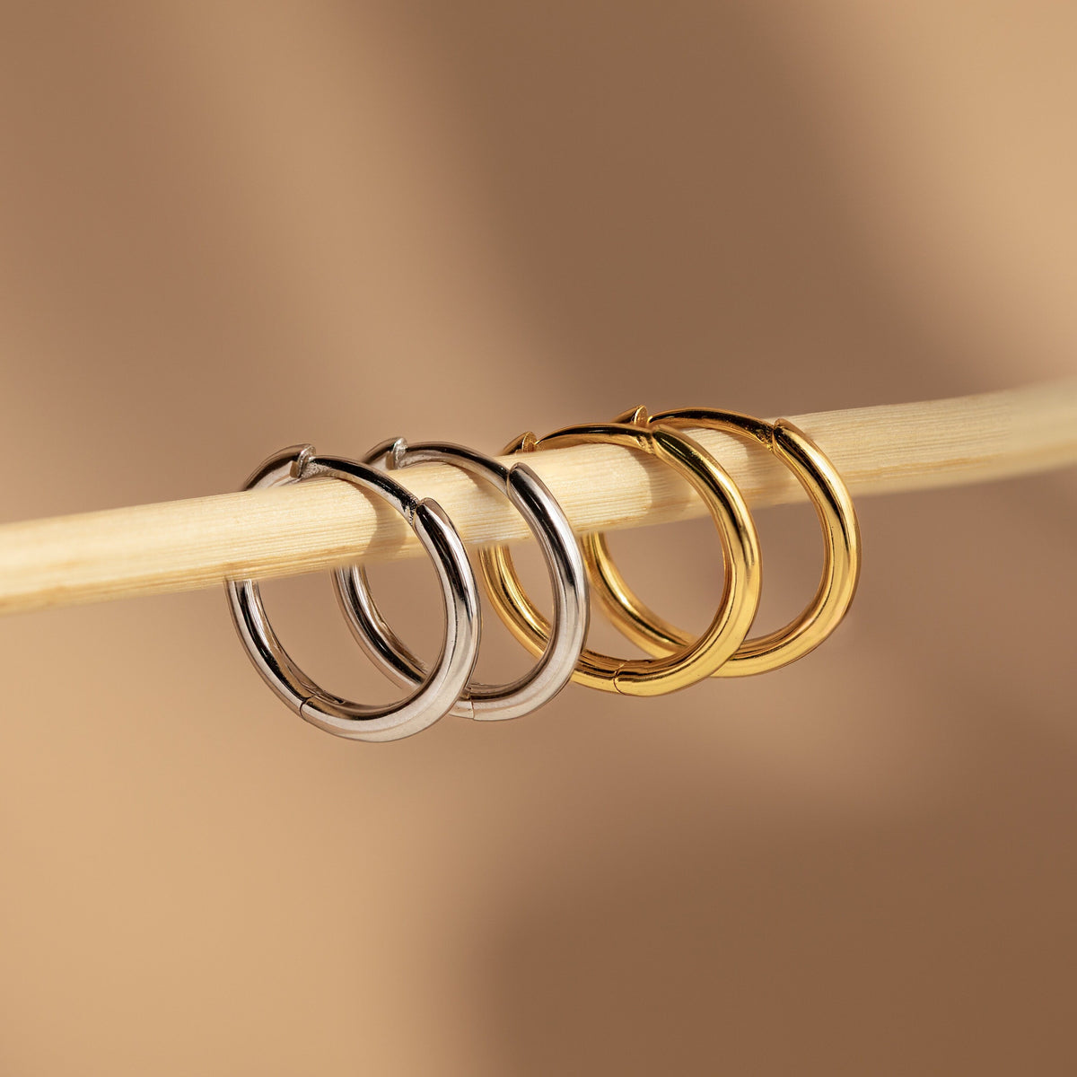 The Mini Bold Hoops Set—featuring one silver and one gold pair—hangs elegantly on a light wooden stick against a beige background, highlighting the timeless beauty of classic hoop earrings.