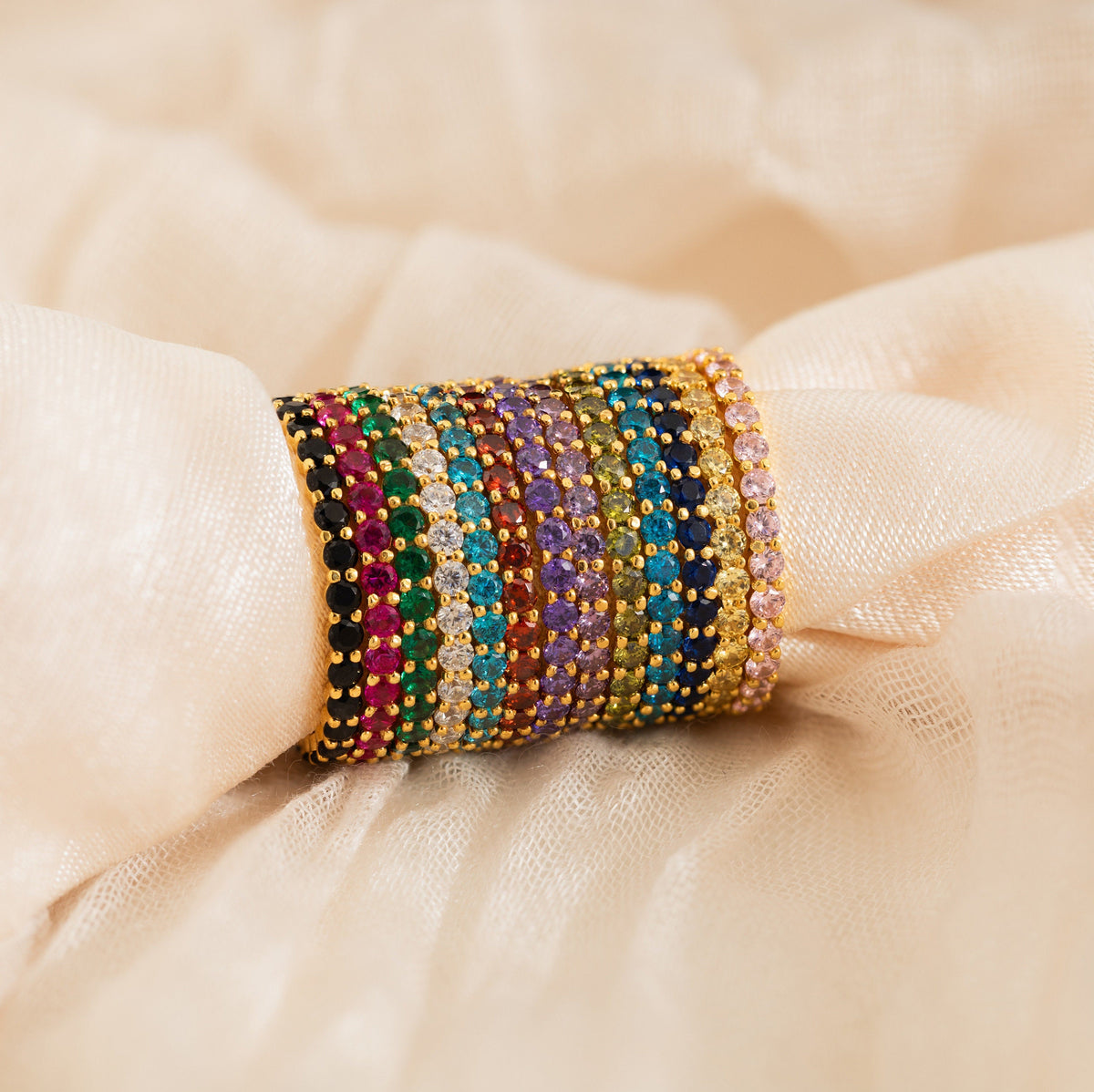 A stack of colorful gemstone-studded gold bangles rests on soft beige fabric, perfect as a Christmas gift or paired with the Eternity Tennis Birthstone Ring for added elegance.