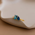 Toi et Moi Birthstone Ring in Rose Gold featuring two blue gemstones—one square, one teardrop—set on a wavy dish. Customizable and perfect as a unique Christmas gift.