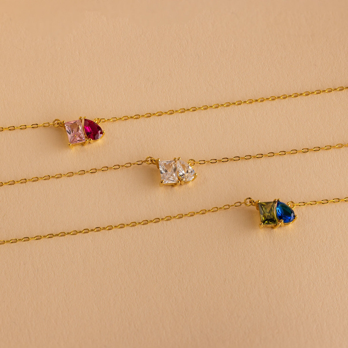 The Toi et Moi Birthstone Necklace features three delicate gold chains, each adorned with two colorful square and teardrop-shaped pendants—an elegant, personalized piece perfect as custom birthstone jewelry.