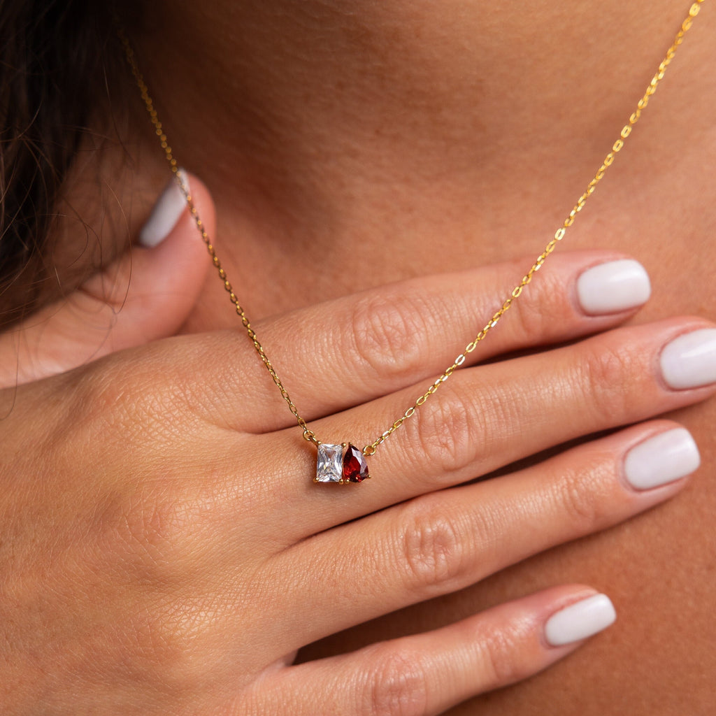 A hand with light pink nails touches the Toi et Moi Birthstone Necklace, which has a gold chain with red and clear gemstone pendants.