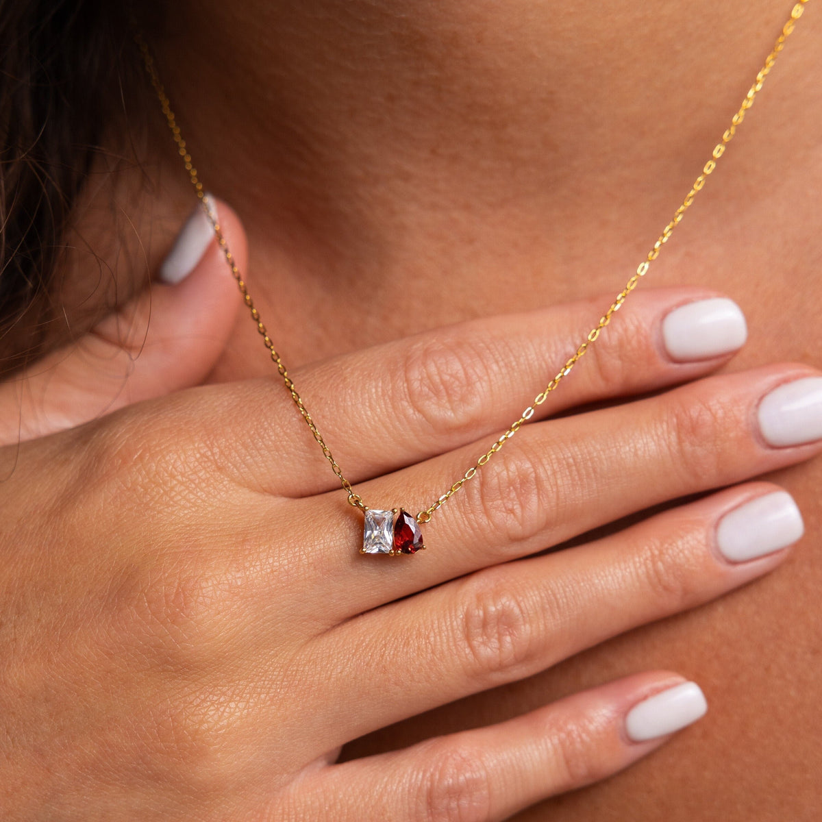 A hand with light pink nails touches the Toi et Moi Birthstone Necklace, which has a gold chain with red and clear gemstone pendants.