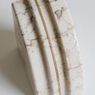 The Marie Chain by LLUME Jewelry, an 18k gold-filled necklace, is displayed draped over a polished marble surface with white, beige, and brown veining and carved grooves.