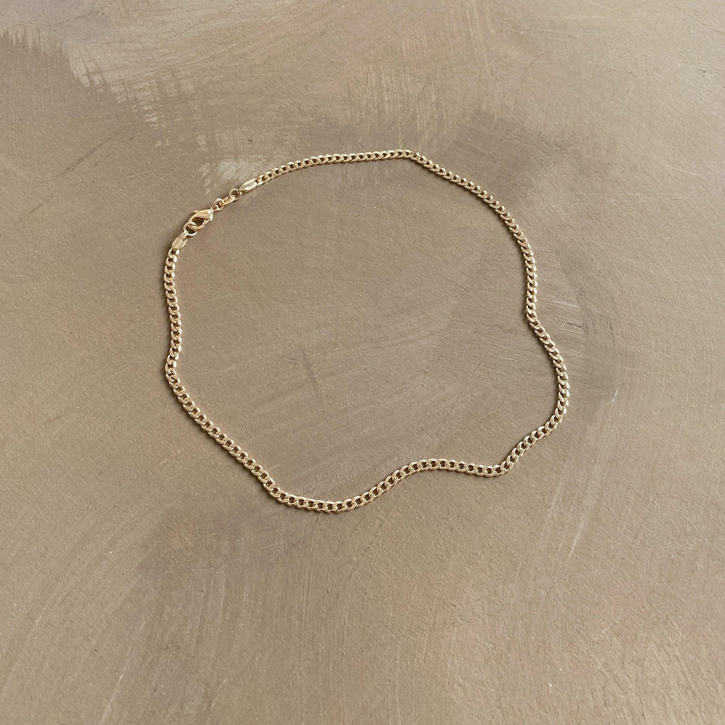 The Marie Chain by LLUME Jewelry, a thin 18k gold-filled necklace with a simple clasp, is displayed in a loose oval shape on a smooth, light brown surface.