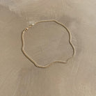 The Marie Chain by LLUME Jewelry, a thin 18k gold-filled necklace with a simple clasp, is displayed in a loose oval shape on a smooth, light brown surface.