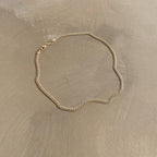 The Marie Chain by LLUME Jewelry, a thin 18k gold-filled necklace with a simple clasp, is displayed in a loose oval shape on a smooth, light brown surface.