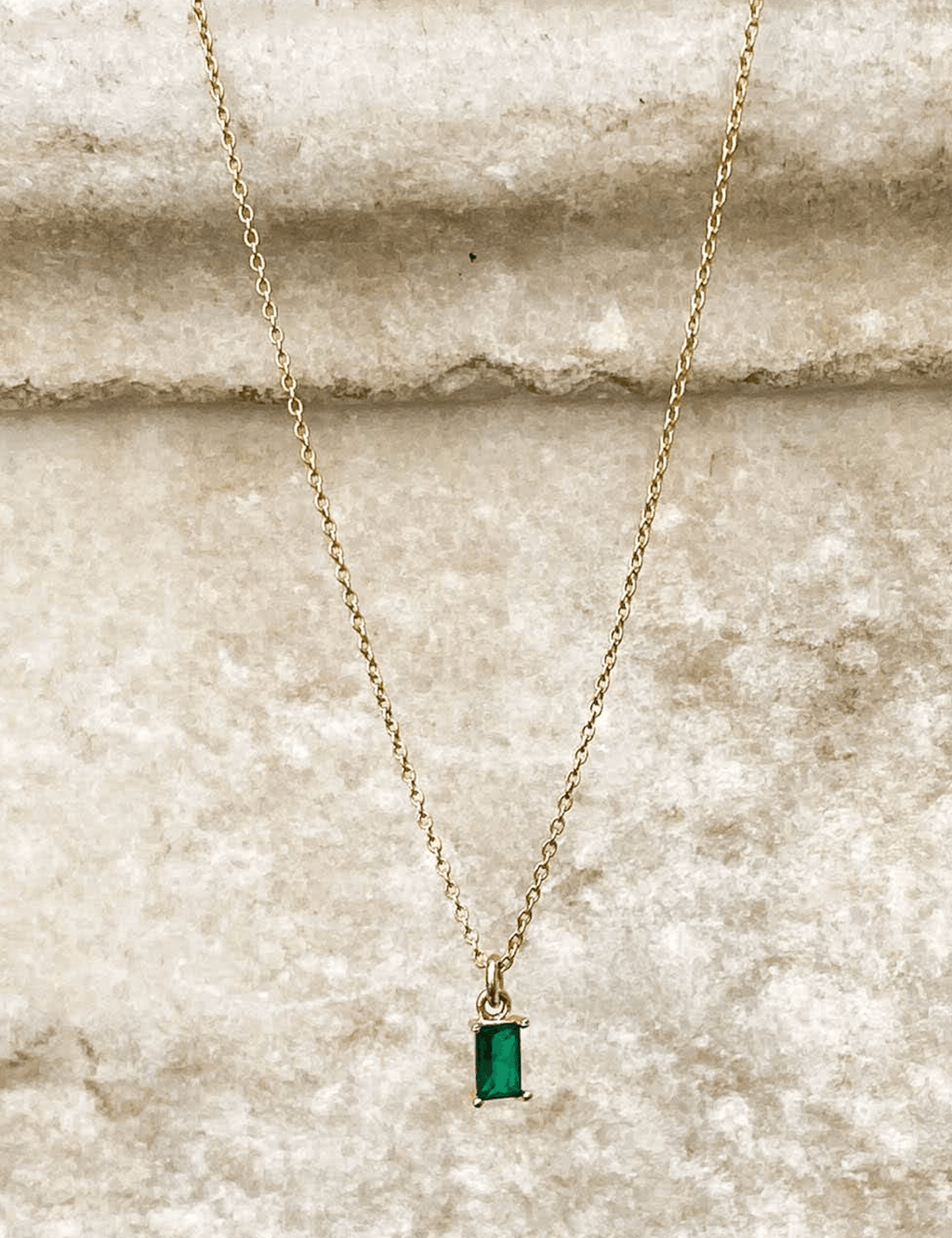 The Kismet Necklace features a gold plated chain with a small rectangular emerald green charm, showcased on a textured beige stone background.