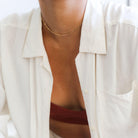 A person wears LLUME Jewelry's Linden Chain, styled with a rust top and white button-up. The photo focuses on their neck and chest, highlighting the subtle elegance of this dainty gold necklace layered for a chic look.