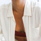 A person wears LLUME Jewelry's Linden Chain, styled with a rust top and white button-up. The photo focuses on their neck and chest, highlighting the subtle elegance of this dainty gold necklace layered for a chic look.