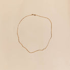 The Harlow Chain by LLUME Jewelry, a delicate 18k gold filled necklace with a subtle twisted design, is arranged in an uneven oval on a light beige background.