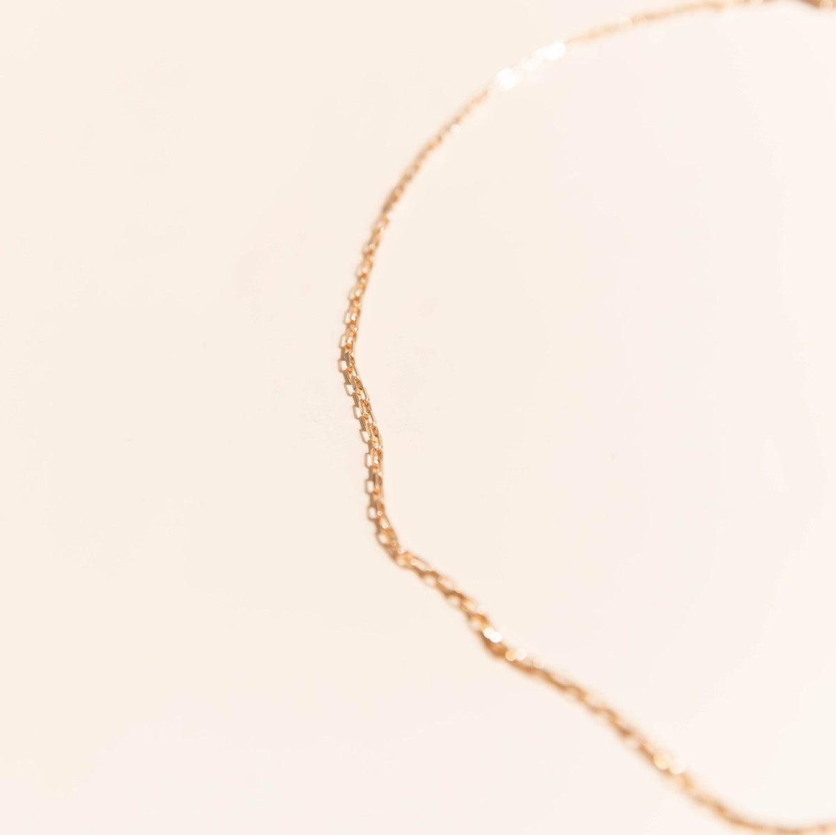 The Harlow Chain by LLUME Jewelry, a delicate 18k gold filled necklace, rests in a gentle curve on a soft beige background with a shallow depth of field softly blurring the chain and backdrop.