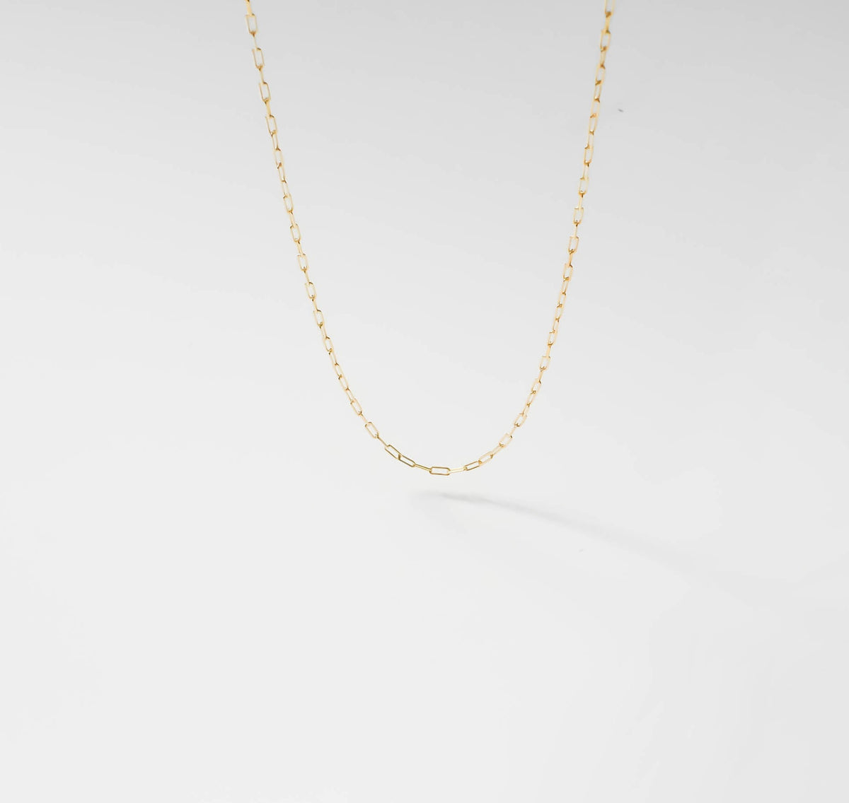 The Adelene Paperclip Chain by LLUME Jewelry features small, oval gold-plated links. Shown against a white background, the adjustable necklace forms a gentle curve and casts a subtle shadow below.
