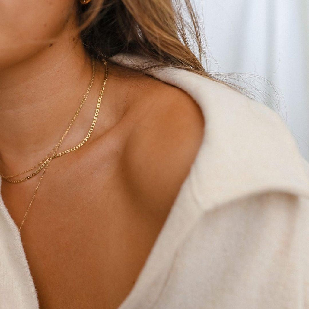 Close-up of a woman in a cream off-shoulder top wearing LLUME Jewelry's waterproof Marie Chain, an 18k gold-filled necklace. Only part of her face, neck, shoulder, and hair are visible against a softly blurred background.