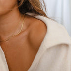 Close-up of a woman in a cream off-shoulder top wearing LLUME Jewelry's waterproof Marie Chain, an 18k gold-filled necklace. Only part of her face, neck, shoulder, and hair are visible against a softly blurred background.