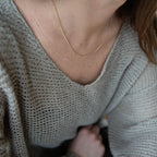 A close-up shows a person wearing a loose-knit light gray sweater and the LLUME Jewelry Linden Chain, an 18K gold plated necklace resting delicately on their neck, with their face hidden and hair softly draped to one side.