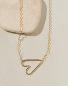 Delicate gold chain necklace with a sideways open-outline gold heart pendant on cream surface with stone pebble