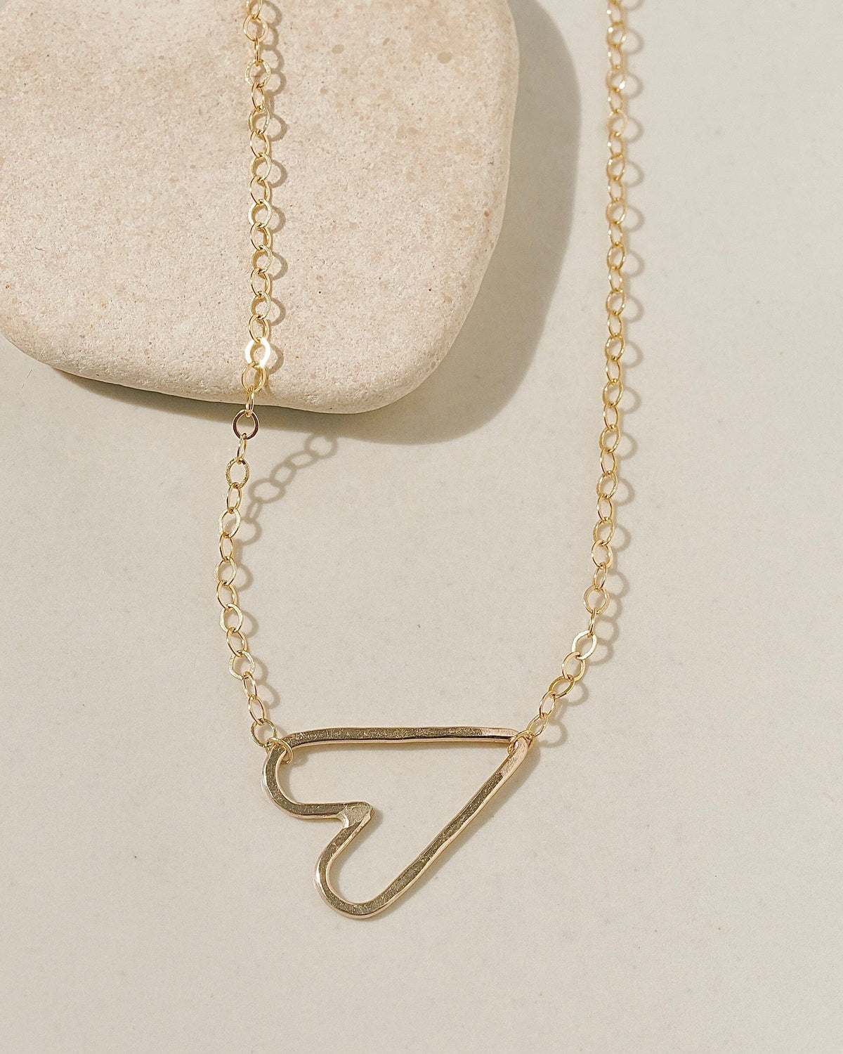 Delicate gold chain necklace with a sideways open-outline gold heart pendant on cream surface with stone pebble