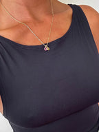 A person wearing a sleeveless black top and the LLUME Jewelry Build a Birthstone Necklace, featuring a gold chain with a small purple birthstone pendant and gold-plated charms, shown from shoulders to upper chest.