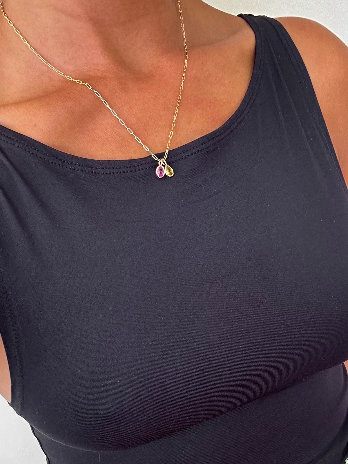 A person wearing a sleeveless black top and the LLUME Jewelry Build a Birthstone Necklace, featuring a gold chain with a small purple birthstone pendant and gold-plated charms, shown from shoulders to upper chest.