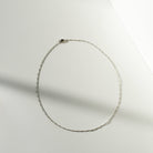 The Colette Necklace Silver by LLUME Jewelry, a delicate sterling silver chain, is arranged in an oval on a white surface with soft light and shadow creating a minimalist background.