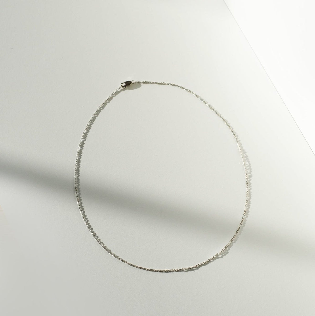 The Colette Necklace Silver by LLUME Jewelry, a delicate sterling silver chain, is arranged in an oval on a white surface with soft light and shadow creating a minimalist background.