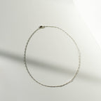 The Colette Necklace Silver by LLUME Jewelry, a delicate sterling silver chain, is arranged in an oval on a white surface with soft light and shadow creating a minimalist background.
