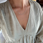 A person wearing a light-colored v-neck long-sleeve blouse is shown from the neck to upper chest, featuring the LLUME Jewelry Colette Necklace Silver—a delicate sterling silver chain.