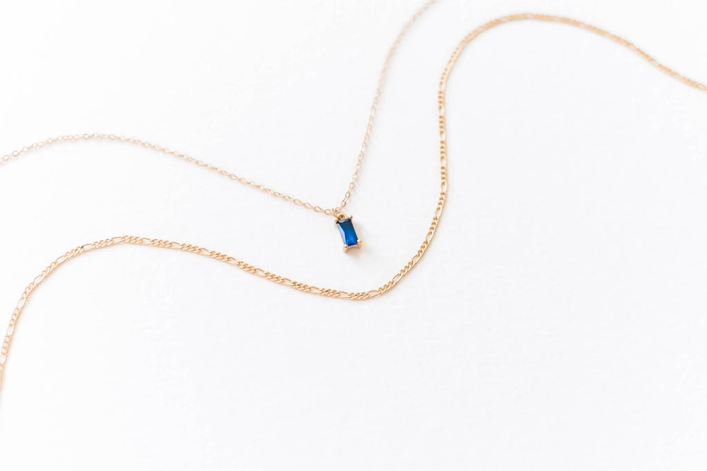 The LLUME Jewelry Taylor Daily Set features a delicate gold pendant necklace with a thin chain and small rectangular blue gemstone, paired with a second slim gold chain—ideal for an elegant layered look.