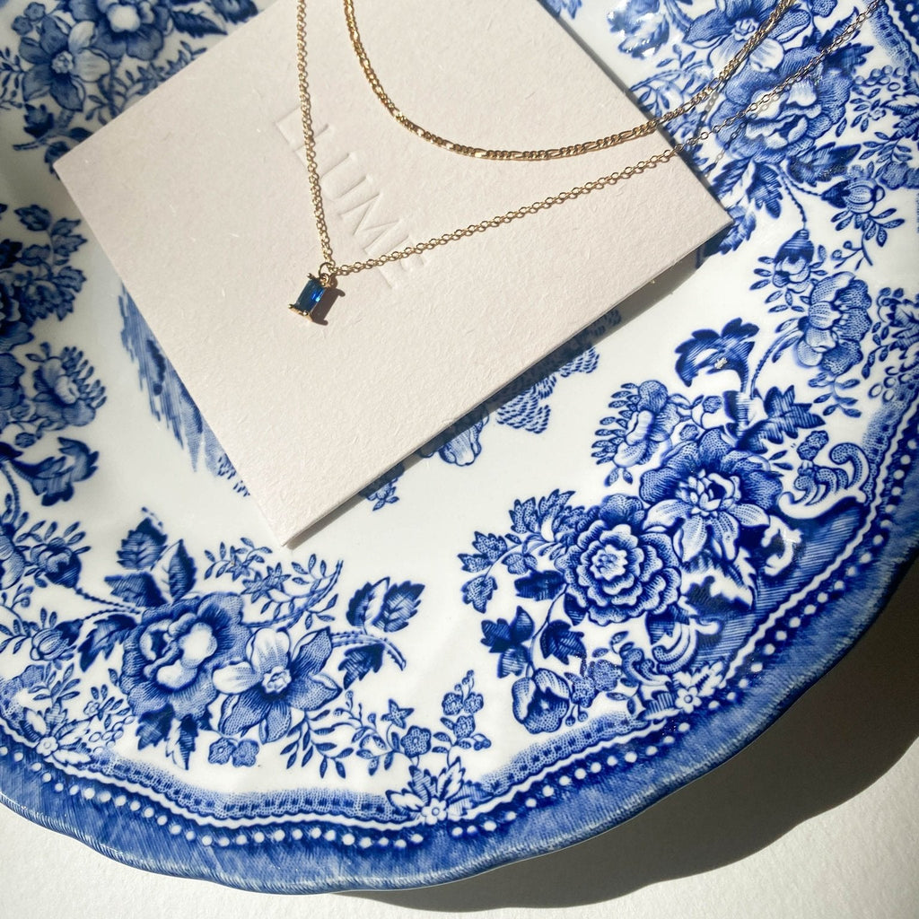 A gold Taylor Daily Set necklace by LLUME Jewelry with a small rectangular blue pendant rests on a white card, displayed on a decorative white and blue floral china plate in bright sunlight.