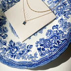 A gold Taylor Daily Set necklace by LLUME Jewelry with a small rectangular blue pendant rests on a white card, displayed on a decorative white and blue floral china plate in bright sunlight.