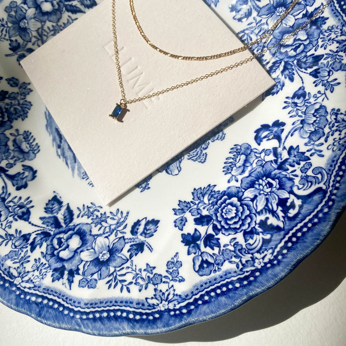 A gold Taylor Daily Set necklace by LLUME Jewelry with a small rectangular blue pendant rests on a white card, displayed on a decorative white and blue floral china plate in bright sunlight.